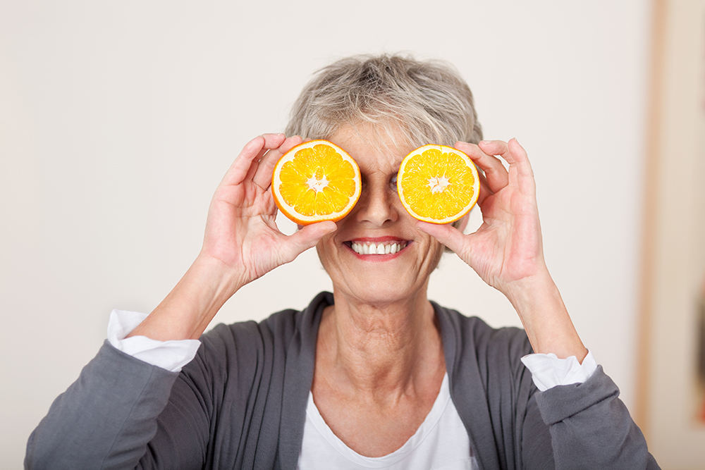 smiling,senior,woman,holding,half,oranges,over,eyes,against,white