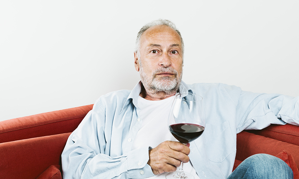 senior,man,sitting,on,couch,holding,wine,glass,with,red